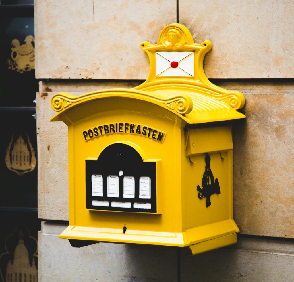 A vintage yellow mailbox against a stone wall with ornate design elements.